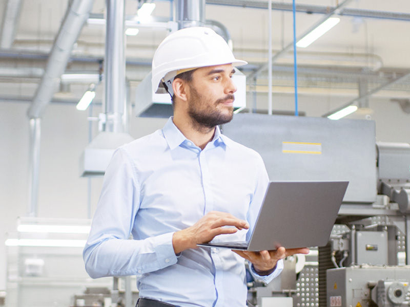 Mann mit Sicherheitshelm und Laptop bei der Kontrolle von Produktionsprozessen in einer Fabrik