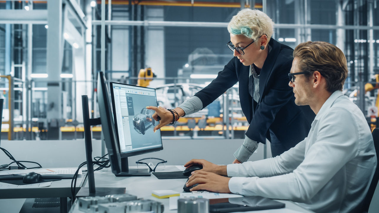 Two automotive engineers discussing concept car blueprints on desktop computer in modern office at assembly plant