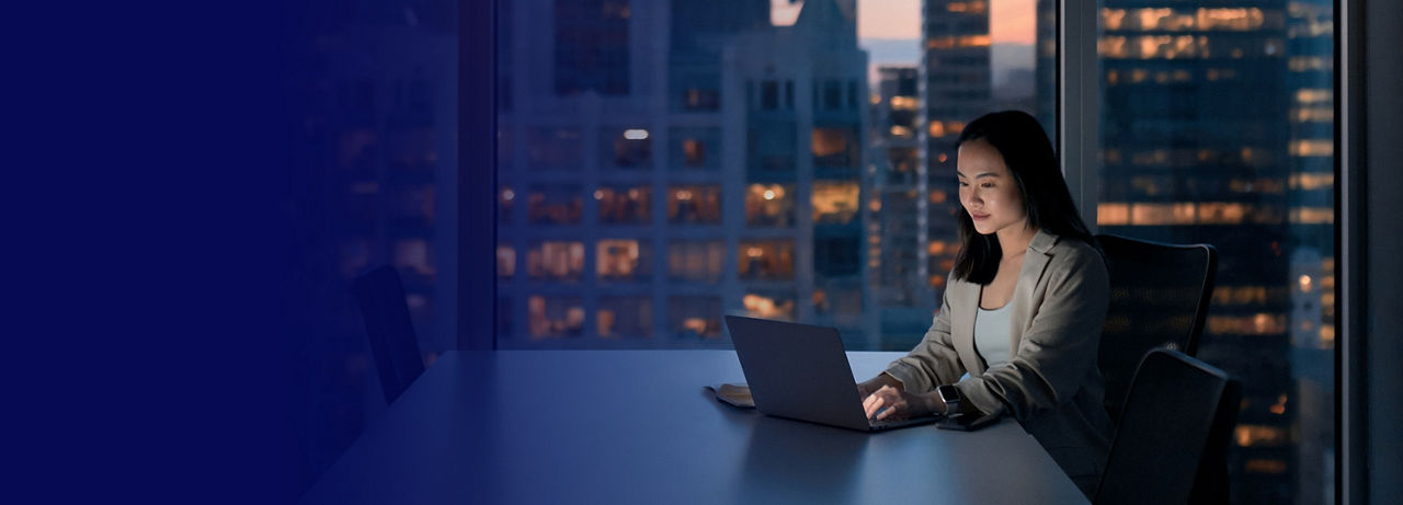 Mujer de negocios asiática joven ocupada trabajando en la computadora portátil en la noche en una oficina corporativa oscura. Mujer de negocios profesional usando su computadora sentada a la mesa, vista de la gran ciudad desde la ventana. Vertical