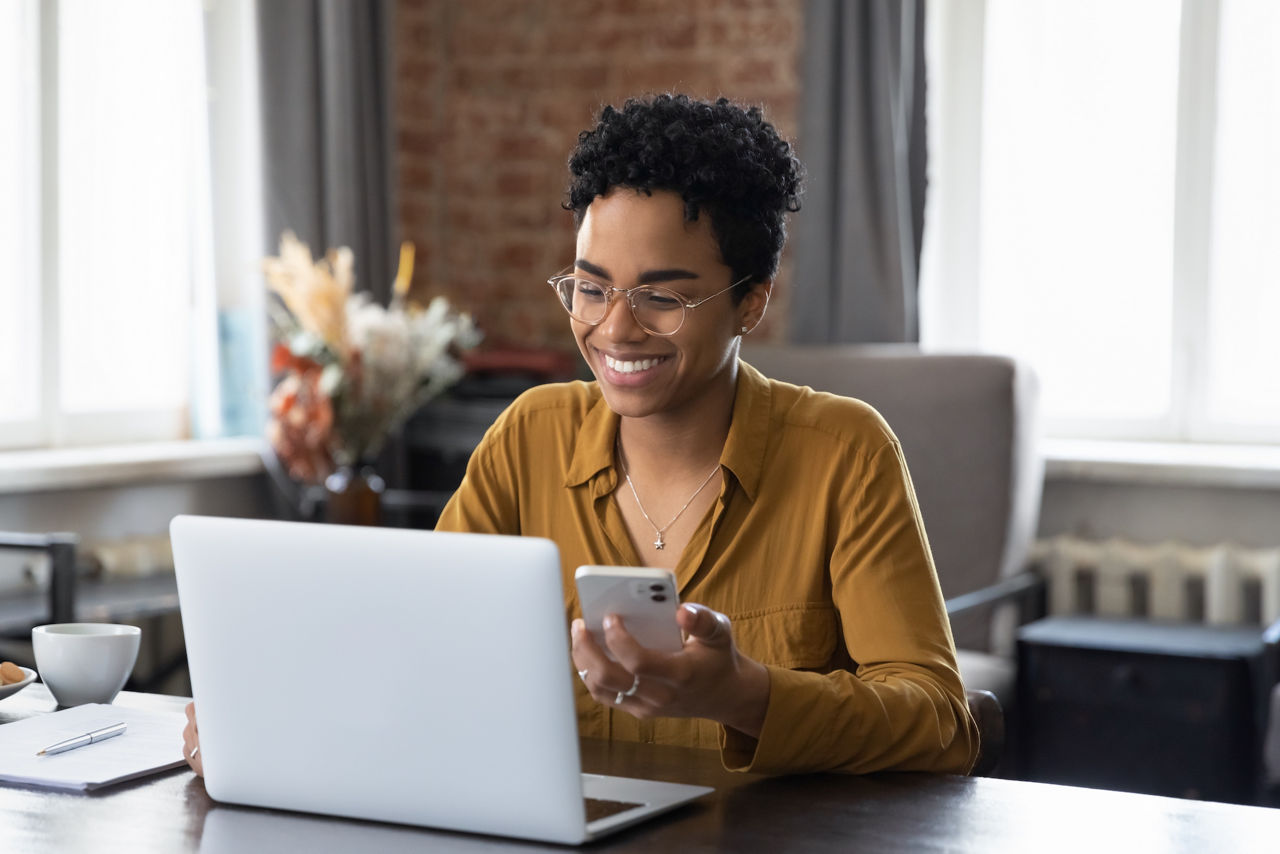 Woman sits at workplace desk holds cellphone staring at laptop