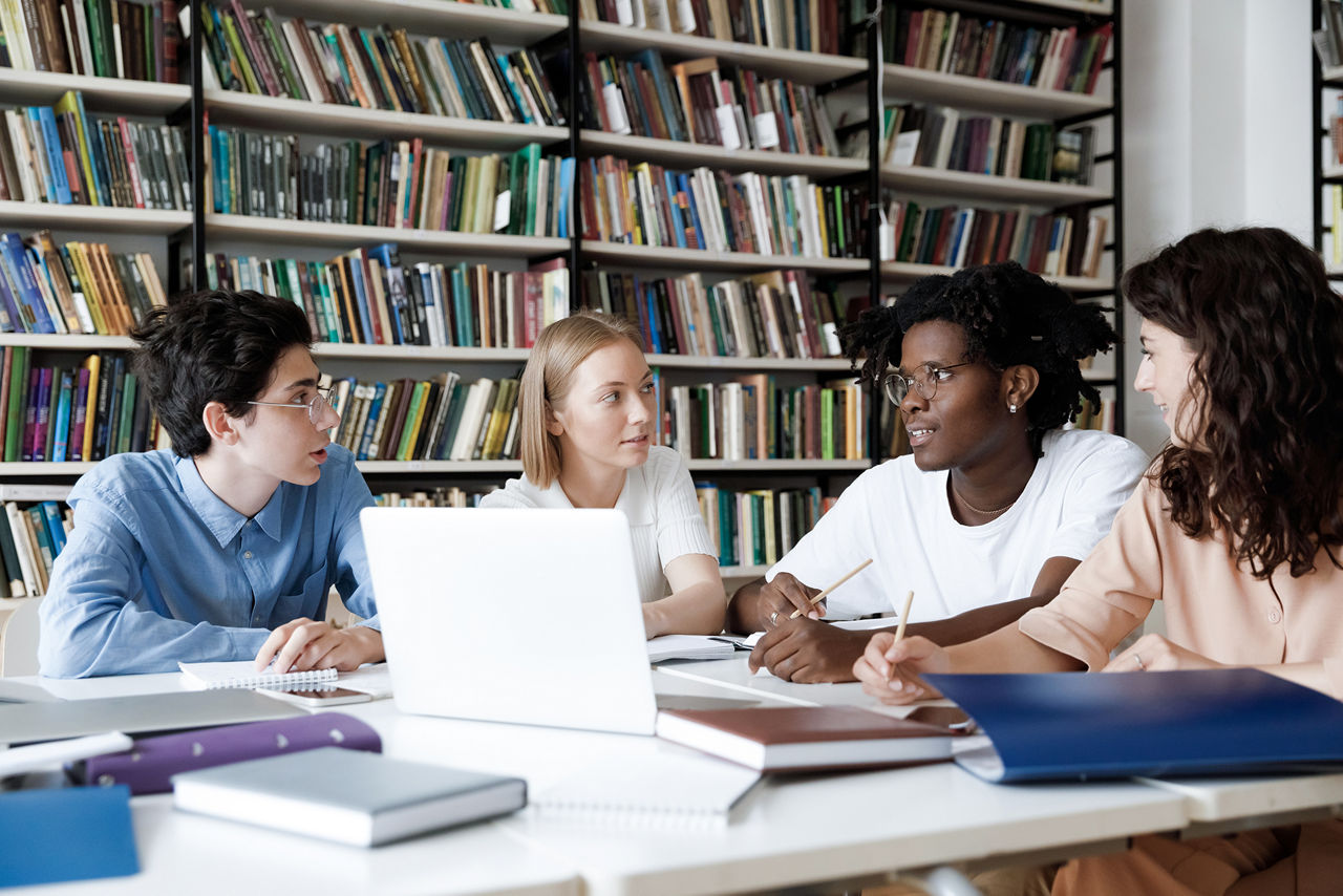 African student guy with schoolmates sit at table in library discuss collaborative task. Multi ethnic schoolgirls schoolboys learn subject, studying together, gain knowledge, prepare for exams concept