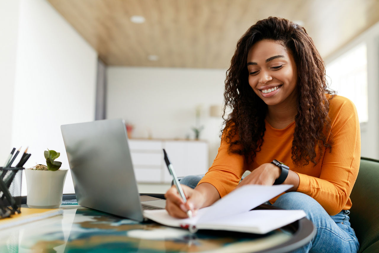 Jovem mulher sorridente sentada à mesa, trabalhando no laptop e escrevendo em um caderno