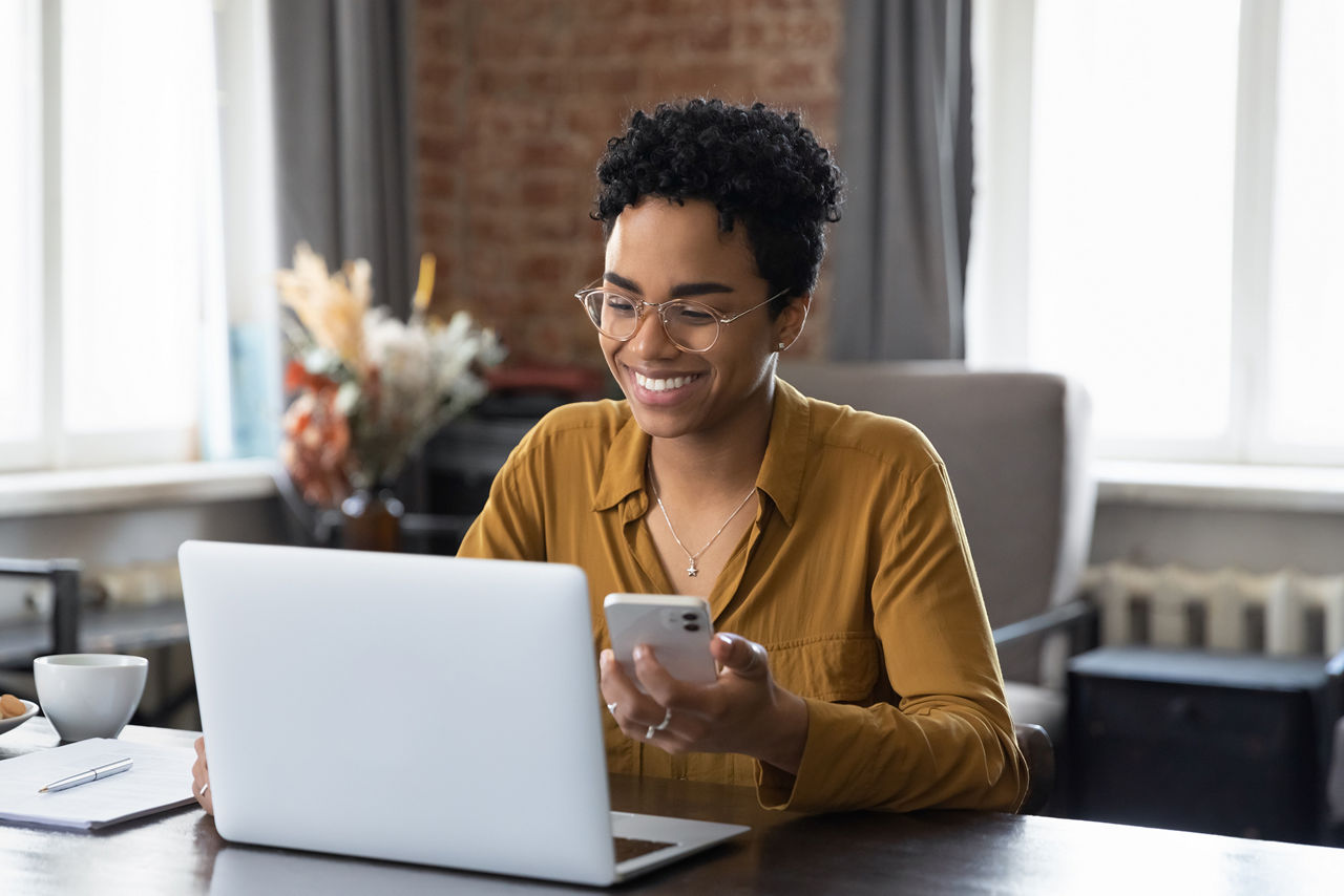 African woman sit at workplace desk holds cellphone staring at laptop, synchronize data between computer and gadget in office, use corporate devices and business application, plan work, use organizer