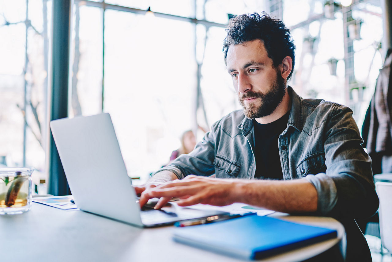 Geconcentreerde jonge man met baard die op afstand aan een project werkt op een moderne laptop met een draadloze internetverbinding
