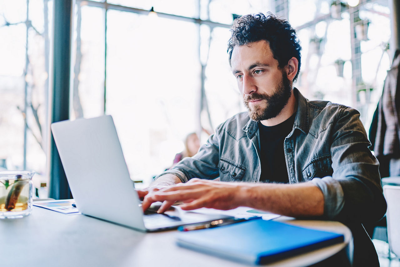 Konzentrierter bärtiger junger Mann, der aus der Ferne an einem Projekt an einem modernen Laptop mit drahtloser Internetverbindung arbeitet