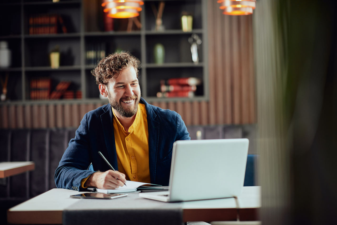 Man writing notes in agenda and looking at laptop while sitting in cafeteria