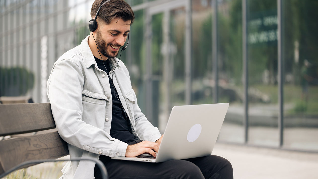 Jonge man op een bank in het park die werkt met headset en laptop