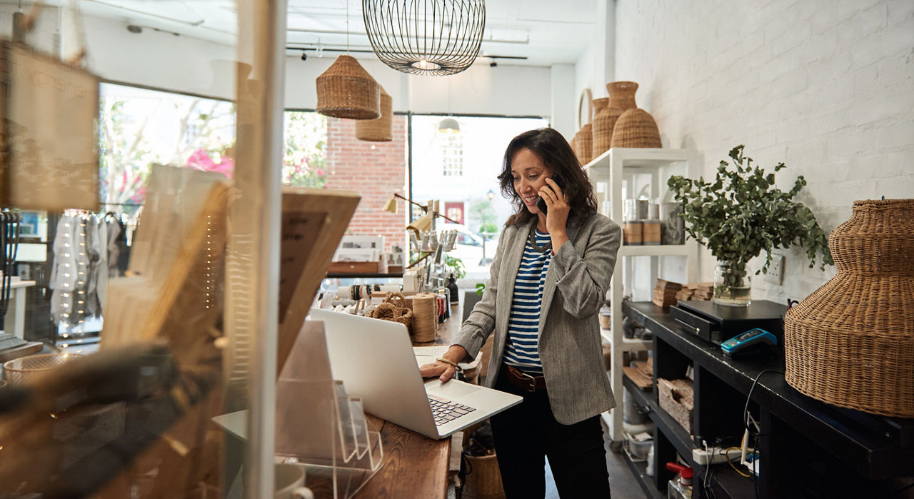 Eine junge Frau steht hinter dem Tresen einer Boutique, arbeitet an ihrem Laptop und telefoniert per Handy