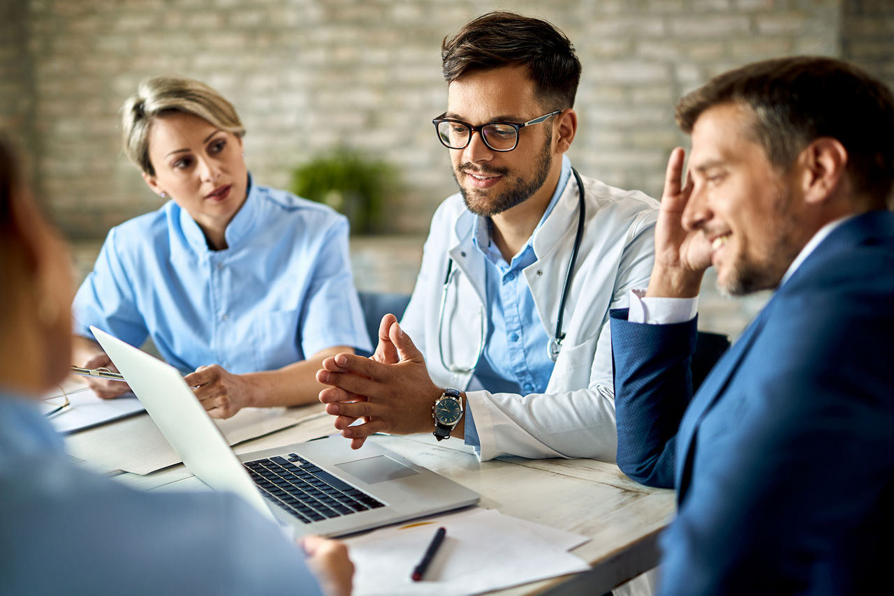 Grupo de profissionais de saúde e um empresário usando um laptop durante uma reunião no escritório. O foco é no jovem médico. 