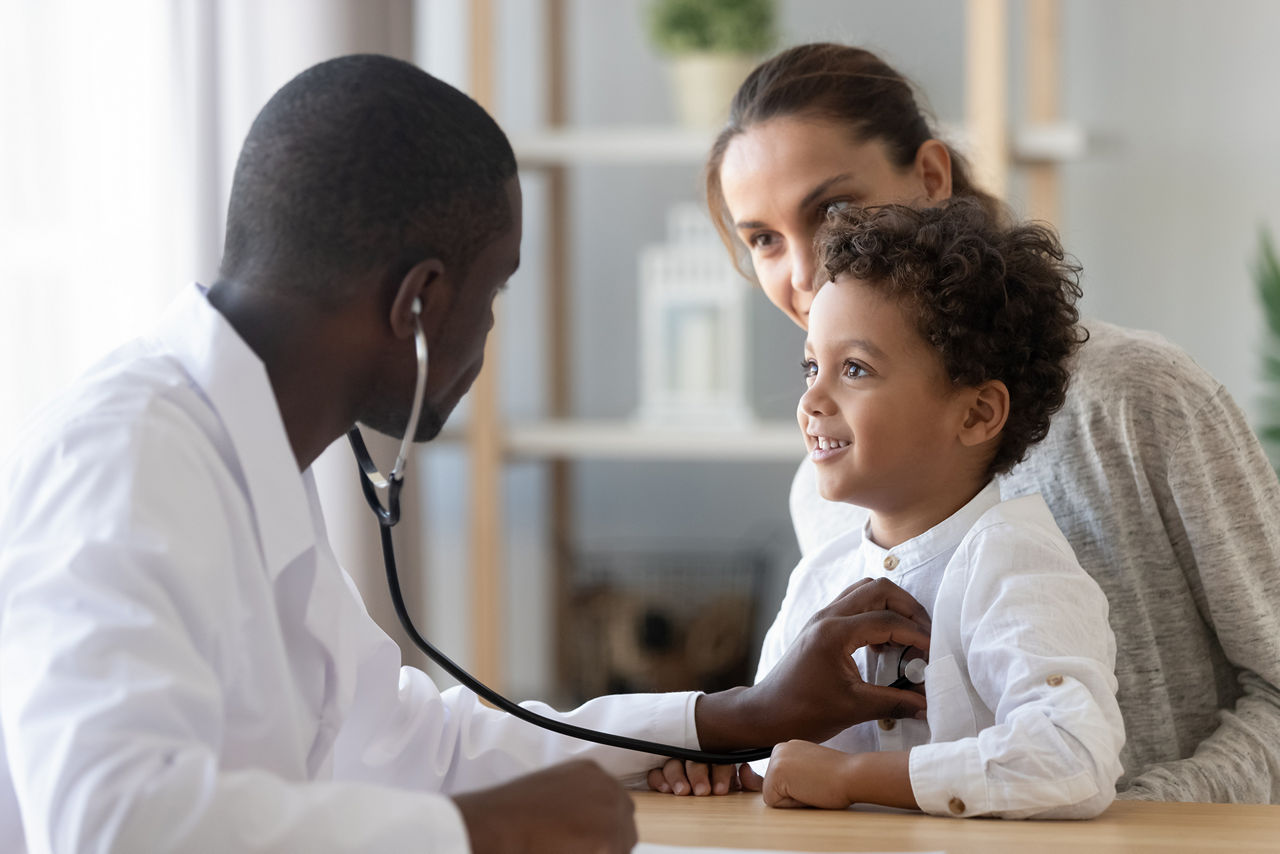 Pédiatre homme muni d’un stéthoscope, écoutant les sons des poumons et du cœur d’un petit garçon assis sur les genoux de sa mère